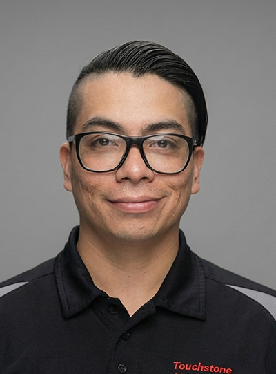 A man with short dark hair combed to the side, wearing black glasses and a black collared shirt with Touchstone embroidered in red, smiles at the camera against a plain gray background.