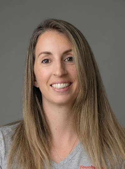 A woman with long, straight, light brown hair smiles at the camera. She is wearing a light gray shirt and is set against a plain gray background.