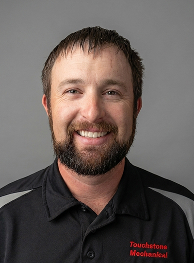 A smiling man with short brown hair and a beard, wearing a black collared shirt with Touchstone Mechanical embroidered in red on the chest, standing in front of a plain gray background.
